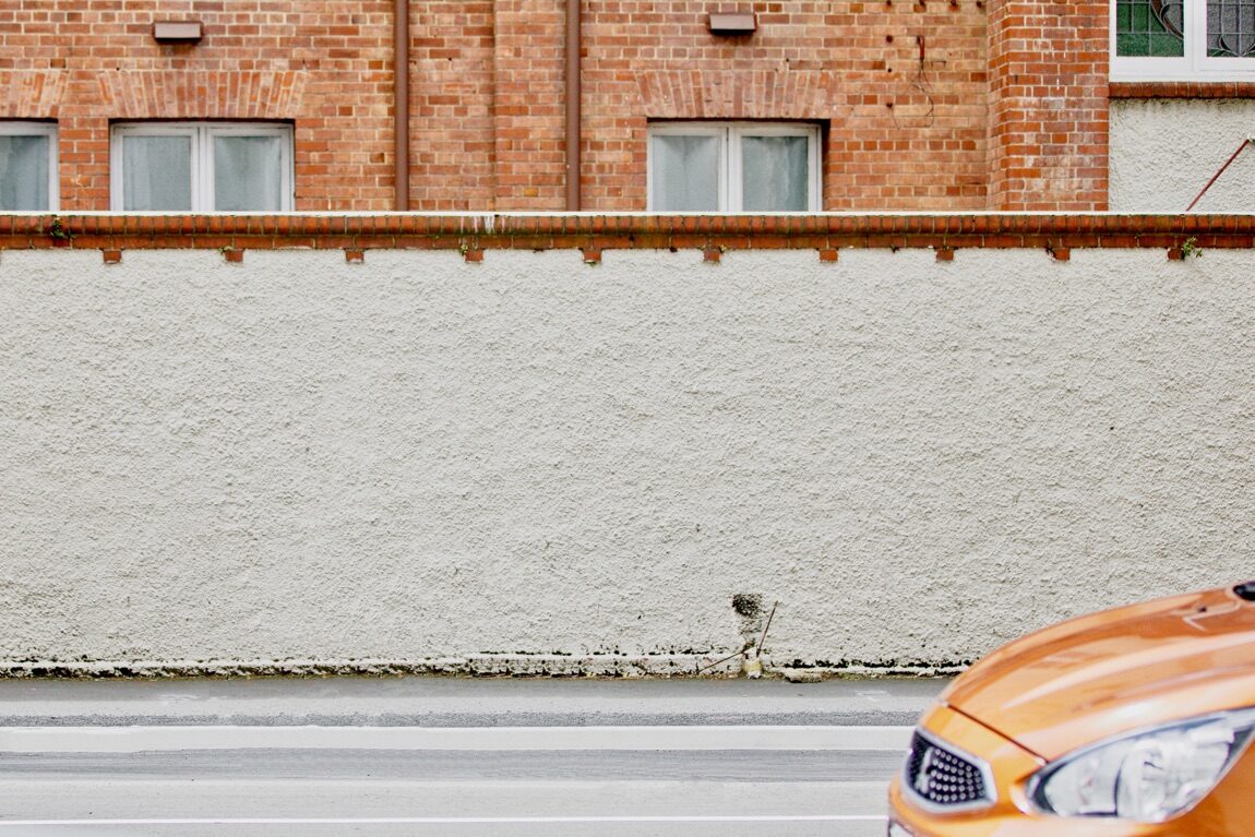 Straße an weißer Mauer, vorne rechts ist die Motorhaube eine Autos zu sehen.