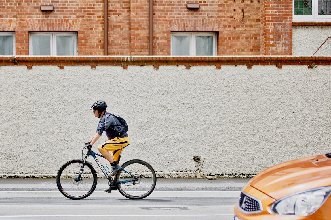 Ein Fahrradfahrer fährt auf der Straße neben einer weißen Mauer. Im Vordergrund rechts ist die Motorhaube eines Autos zu sehen.